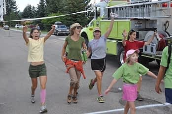 Teton Valley Non-Profits, A jubilant crowd, embodying the spirit of unity and giving fostered by the Community Foundation of Teton Valley, parades past a vibrant green fire truck along a bustling street. Their raised arms and exuberant expressions reflect not just personal joy but also the collective pride in their community's achievements. The lush trees lining the background serve as a testament to their shared commitment to nurturing both nature and neighbor. Through charitable giving, this dedicated organization has empowered local nonprofits and businesses, creating an environment where generosity blossoms into tangible support and growth for all. The Tin Cup Challenge & Run is the CFTV annual giving event day.